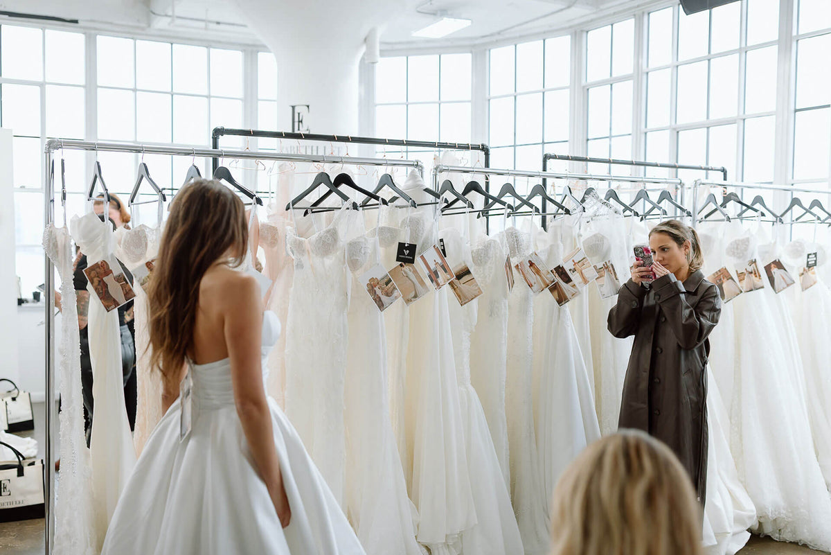 Woman in a white wedding dress standing among hanging gowns in a bridal market.