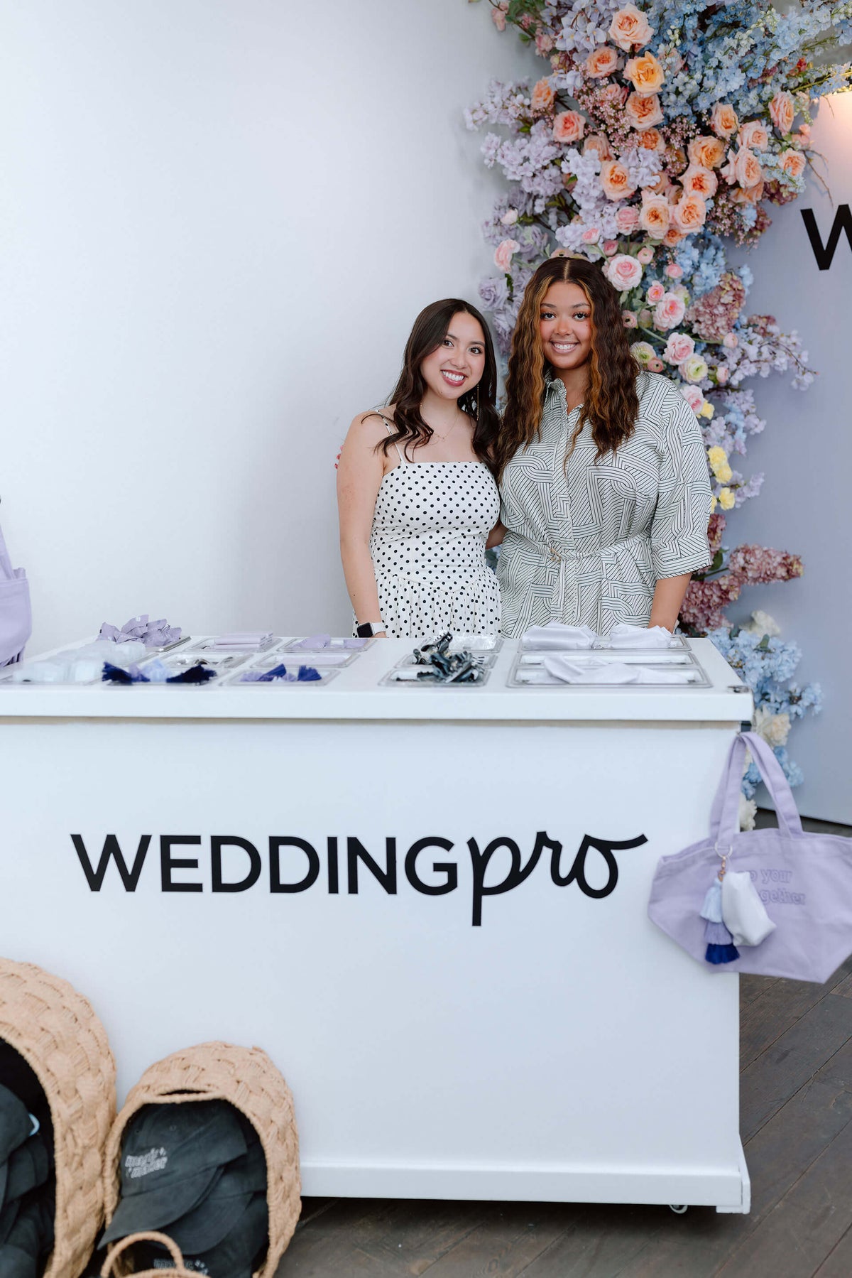 Two women standing behind a the WeddingPro lounge counter with floral decorations in the background.