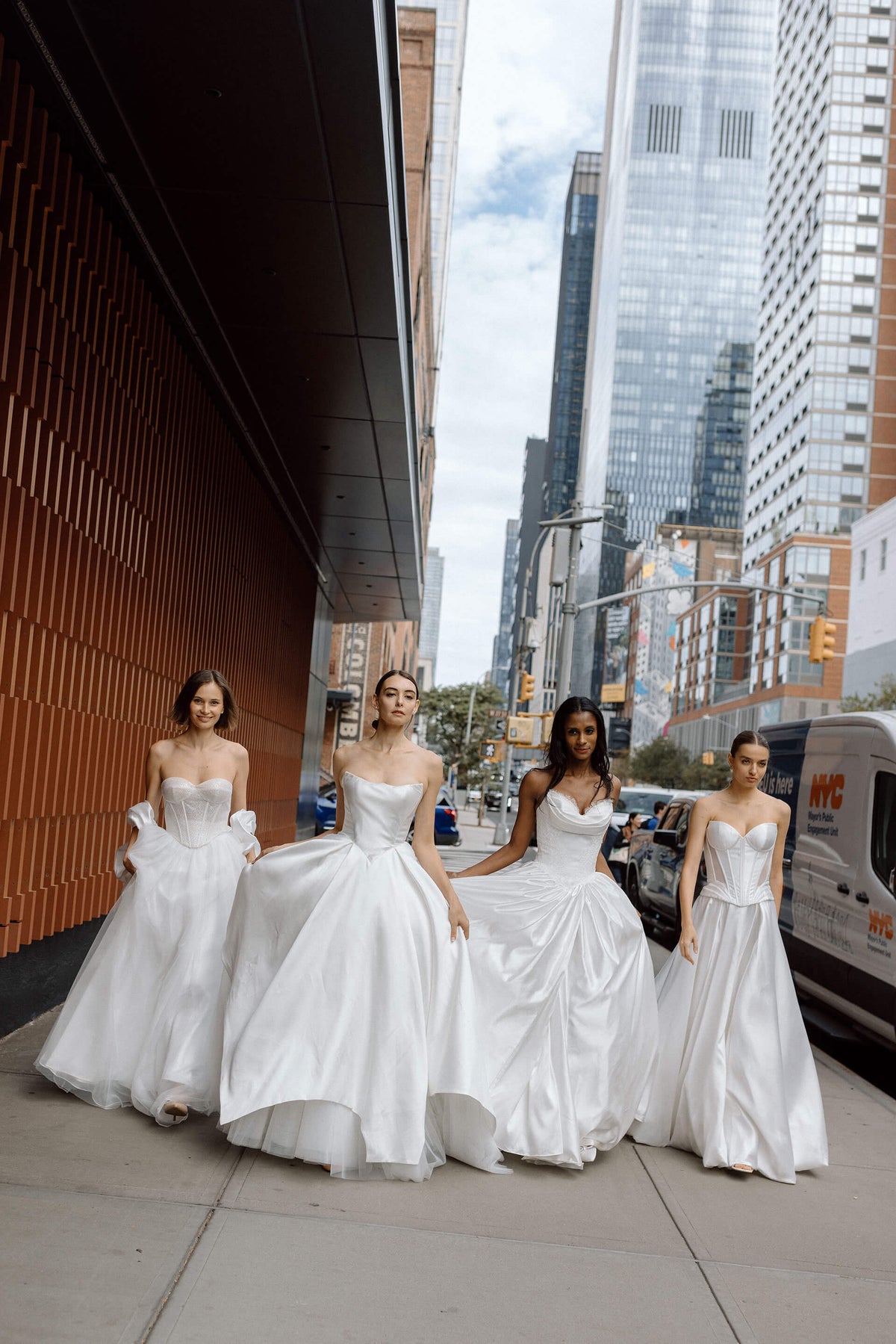Four women in white wedding dresses standing on a city street with tall buildings in the background during the new york bridal fashion week