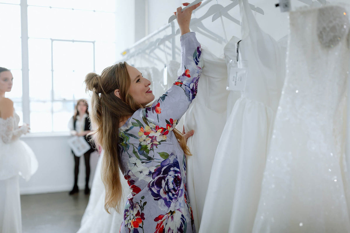 Woman looking at a wedding dress in a store with other dresses and people in the background.