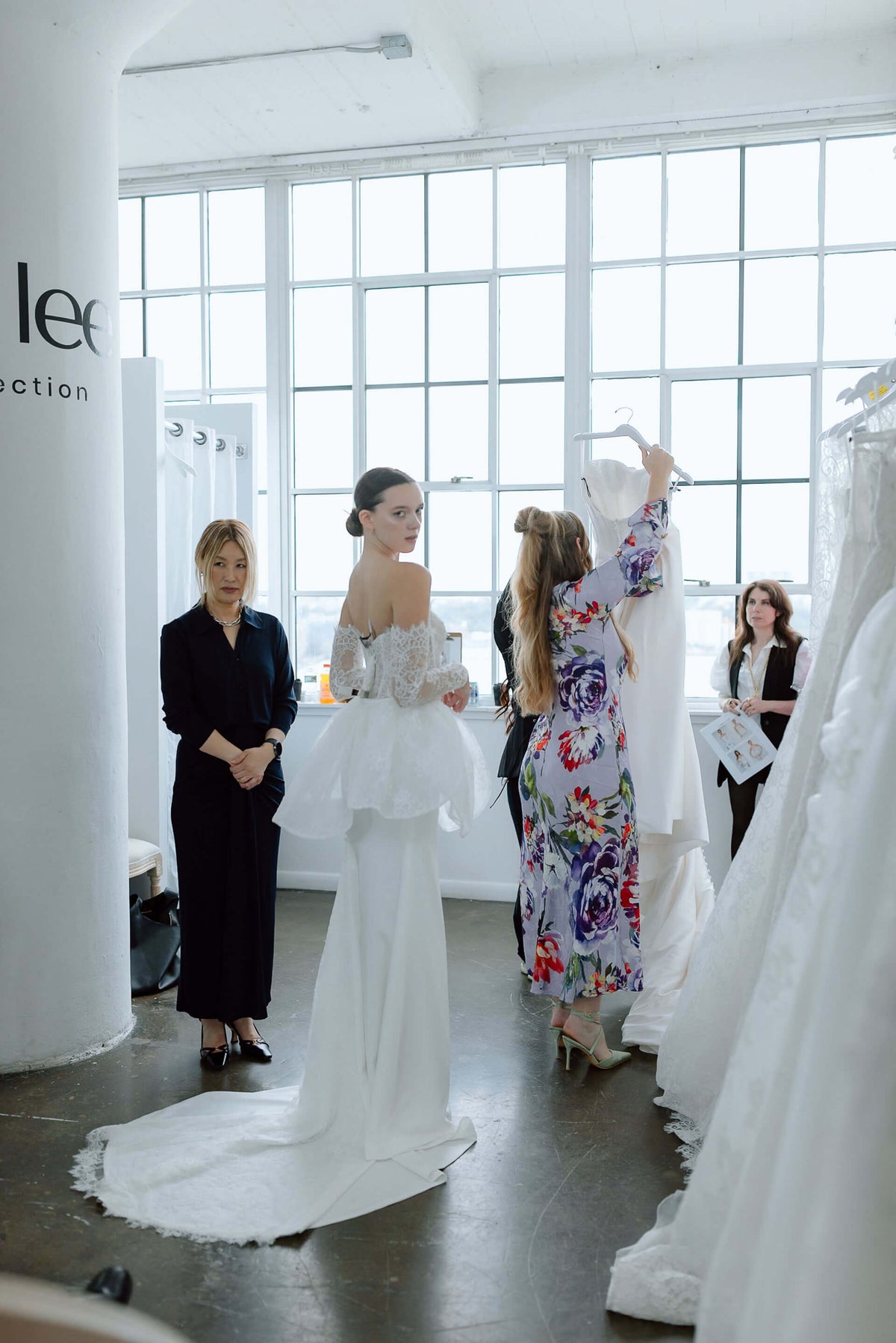 Woman in a white wedding dress standing in a room with large windows and other bridal buyers