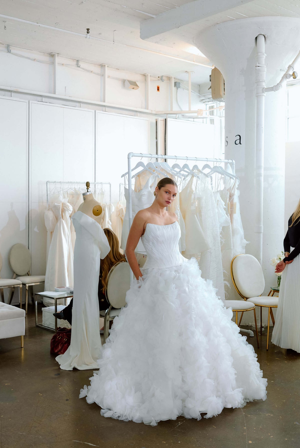 Woman in a white wedding dress standing in a room with other wedding dresses during New York Bridal Fashion Week