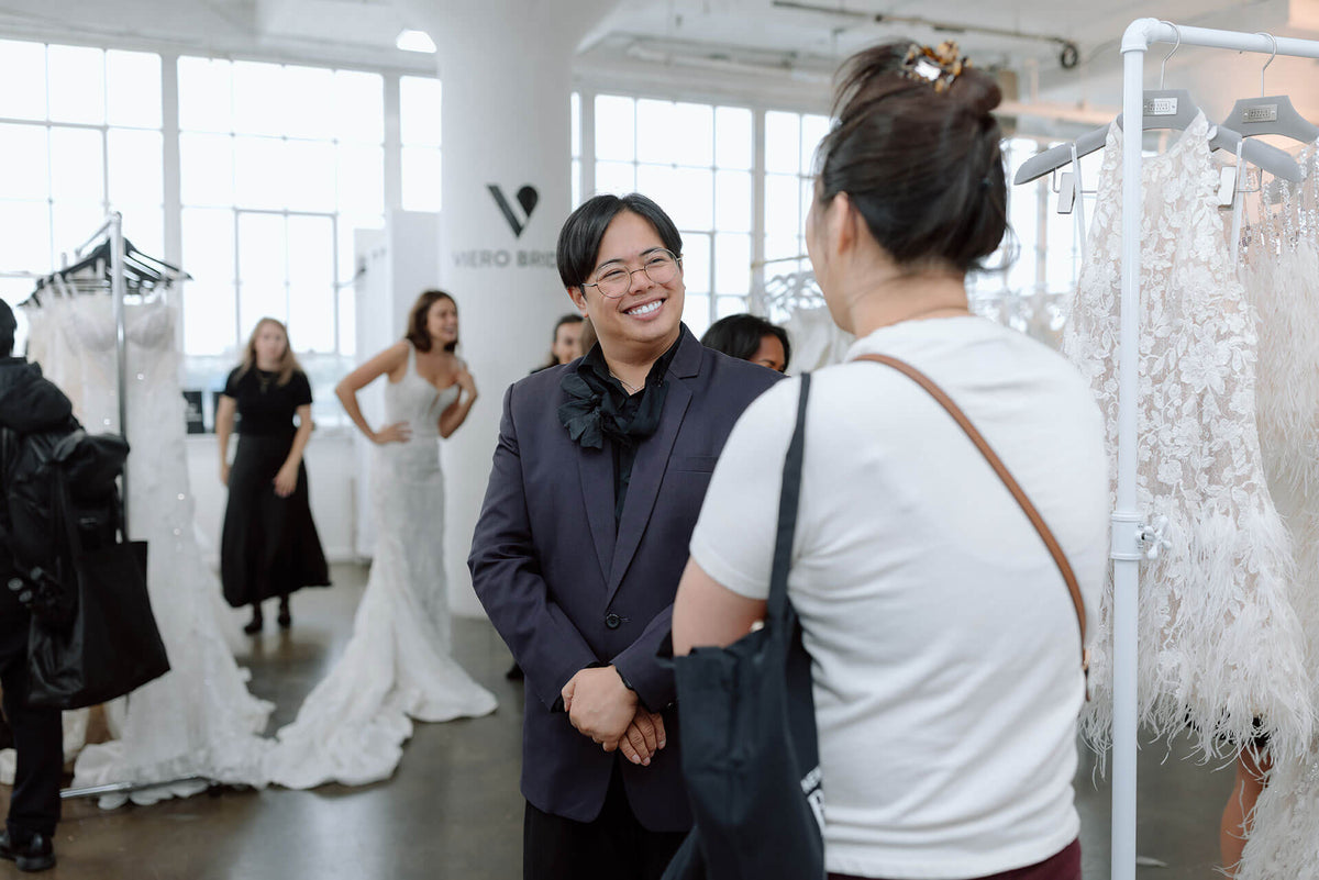Man in a suit standing in a bridal trade show with a woman, surrounded by wedding dresses.